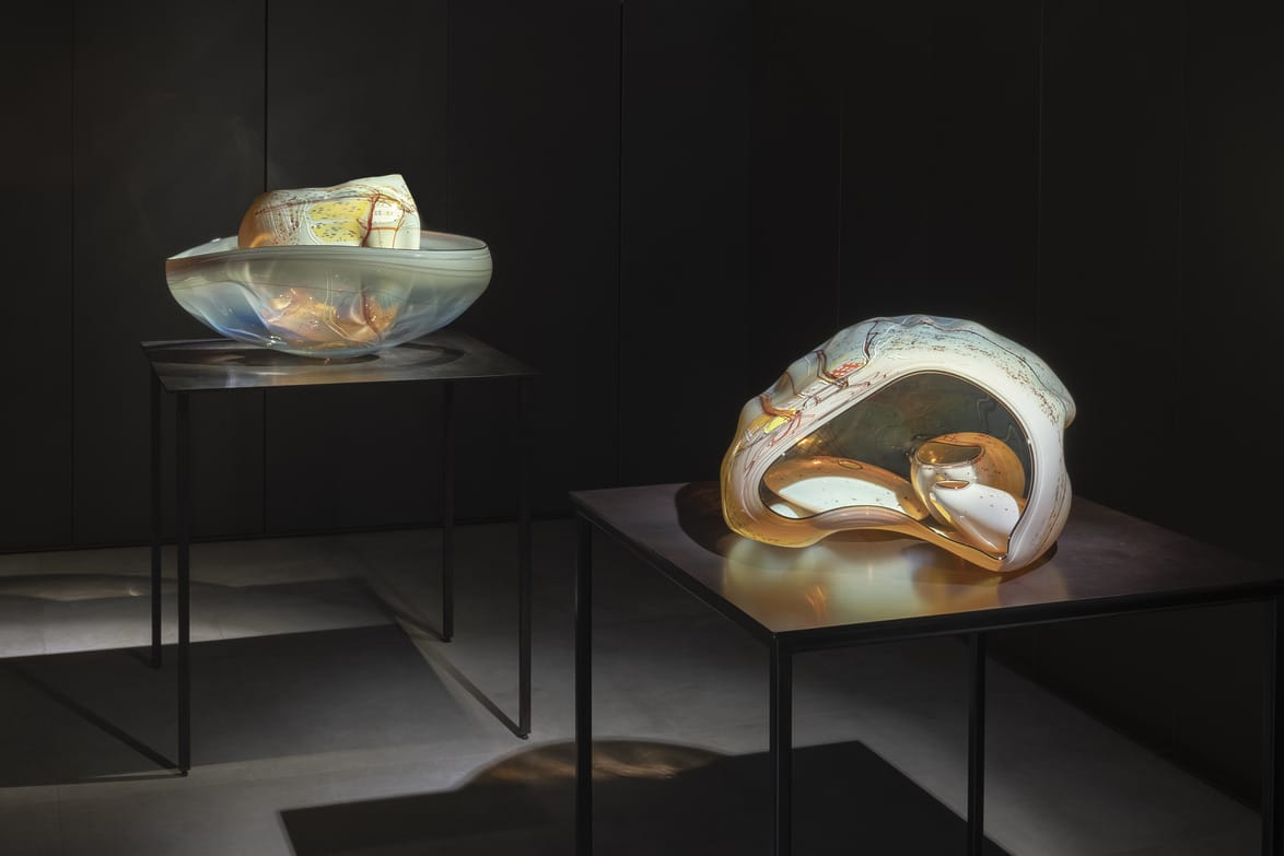 Two abstract glass sculptures on dark metal stands, illuminated in a dimly lit room with reflective surfaces.