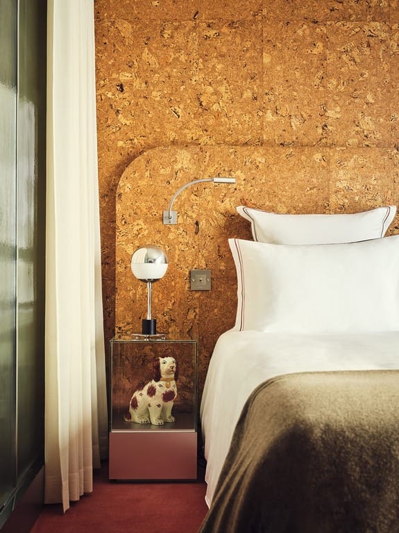 Bedroom with cork wall, white bed, modern lamp on nightstand, and dog figurine in glass display case.