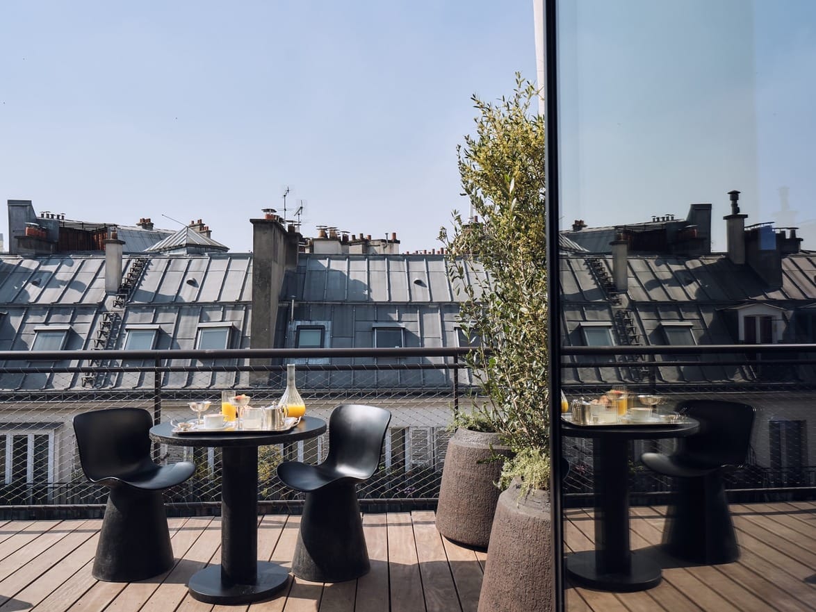 Outdoor balcony with a round table set for breakfast, two black chairs, potted plants, and a rooftop city view.
