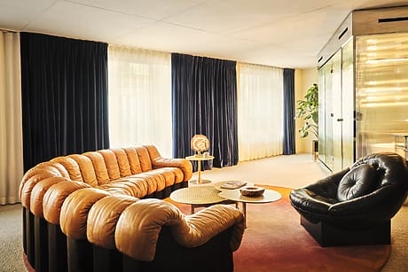 Modern living room with brown tufted sofa, black chair, round coffee tables, dark drapes, and large window illuminating space.