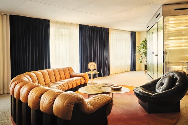 Modern living room with brown tufted sofa, black chair, round coffee tables, dark drapes, and large window illuminating space.