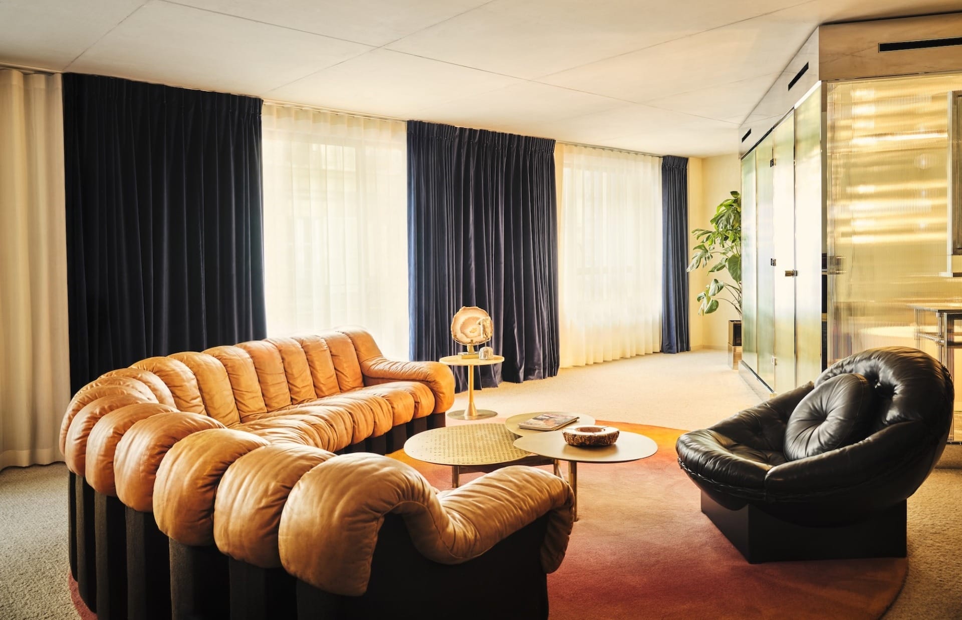 Modern living room with brown tufted sofa, black chair, round coffee tables, dark drapes, and large window illuminating space.