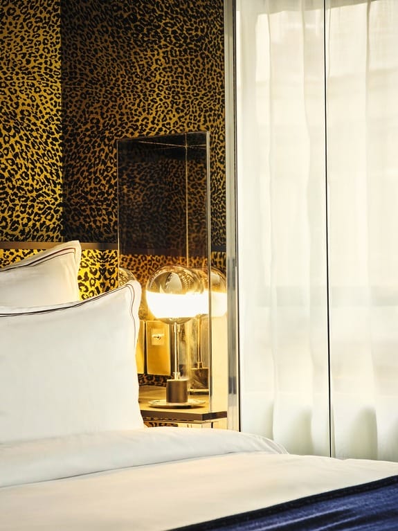 Patterned wallpaper and soft white bedding illuminated by a modern lamp next to a sheer curtain.