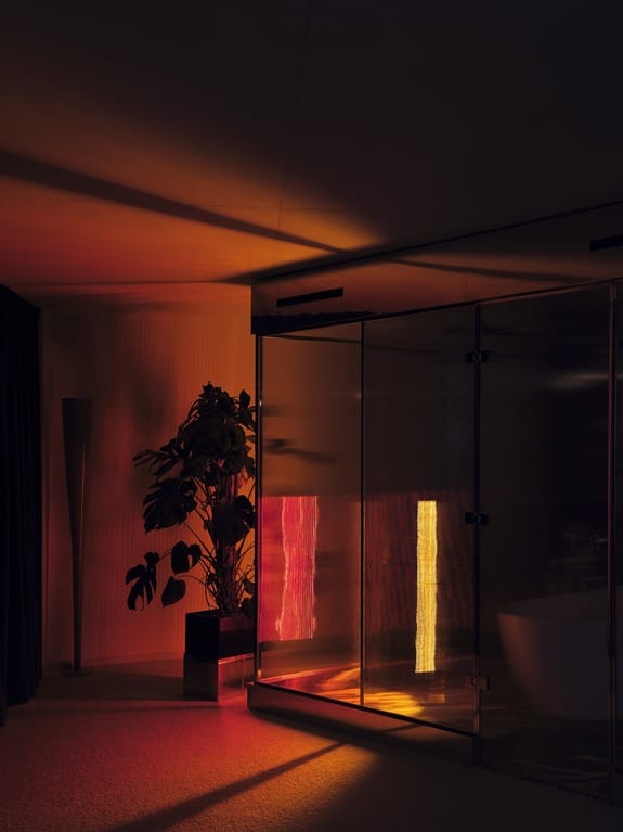 Modern bathroom with glass shower, illuminated by warm, ambient orange and yellow lighting, reflecting off floor and walls.