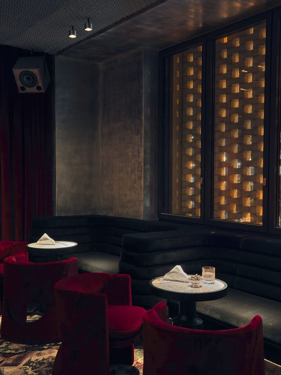 Cozy dimly lit lounge with red chairs, black velvet couches, small tables, and ambient window lighting.