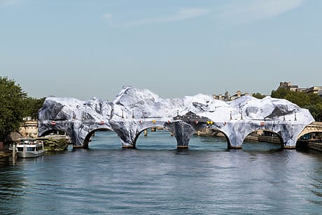 Bridge with stone arches covered in large white and gray fabric resembling mountains, spanning a river with a boat nearby.