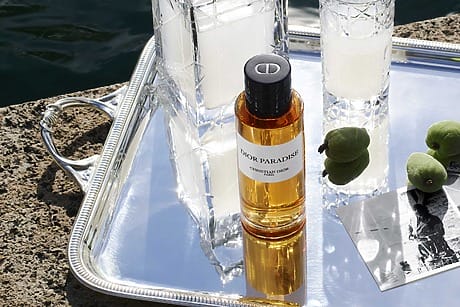 Perfume bottle labeled "Dior Paradise" on silver tray with glasses, green fruits, and photograph nearby.