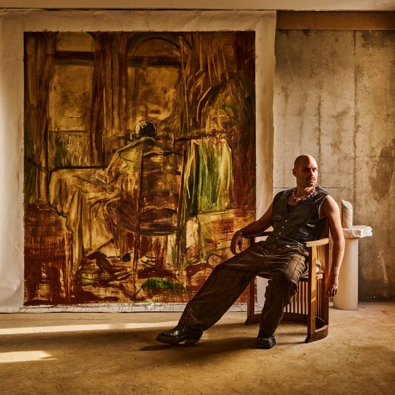 Man sitting on a chair in a room with a large painting on the wall, illuminated by sunlight from a nearby window.