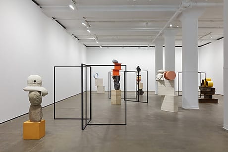 Contemporary art gallery with abstract sculptures and geometric forms displayed on pedestals in a white-walled room.