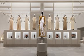 Exhibit of elegant dresses on mannequins in a museum, featuring a central gold piece with historical pottery displayed below at the Costume Institute galleries at the Metropolitan Museum of Art.