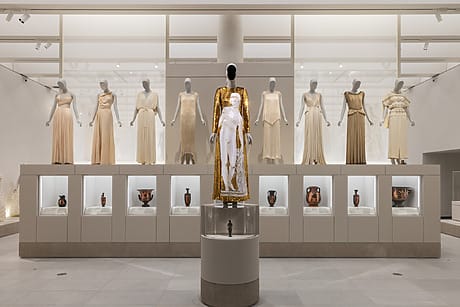 Exhibit of elegant dresses on mannequins in a museum, featuring a central gold piece with historical pottery displayed below at the Costume Institute galleries at the Metropolitan Museum of Art.