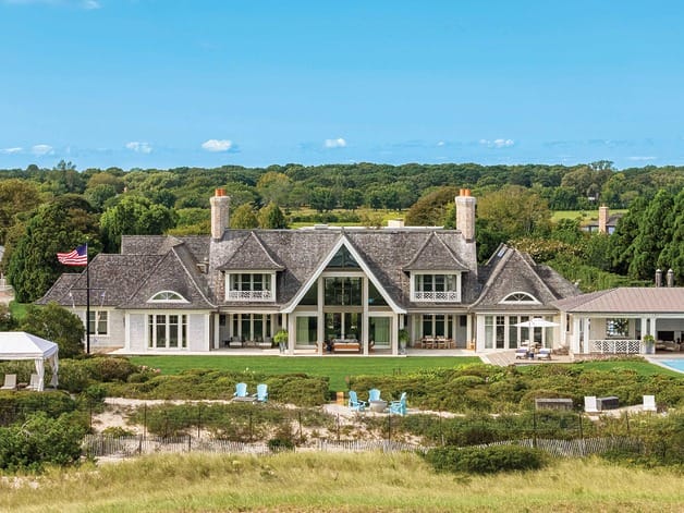 A large modern mansion with a sloped roof, chimneys, and surrounded by greenery with a clear blue sky above.