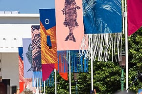 Colorful banners with fish designs and fringe hanging outdoors against a blue sky and greenery in the background.