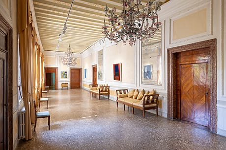 Spacious, elegant hallway with chandeliers, yellow curtains, and cushioned benches in a historic building.