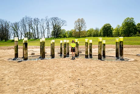 Artist Anicka YI standing among tall, cylindrical stone pillars on a dirt ground with trees in the background.