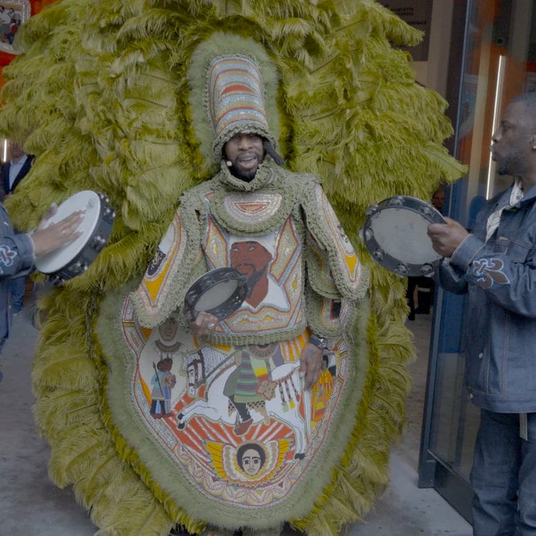 Person in elaborate feathered costume with intricate beadwork, flanked by two individuals holding tambourines.