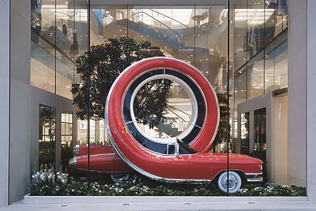A vintage red car displayed inside a unique glass showcase with large circular red sculpture enveloping it.