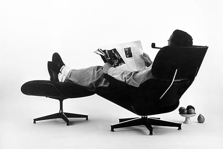 Person reclined in the Eames Lounge, a mid-century modern chair, reading a newspaper with a pipe, beside a small stand with fruit.