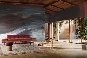 Modern living room with abstract wall mural, wooden ceiling, red sofa, armchair, plants, and large glass doors.