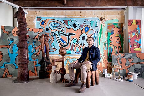 Artist seated in colorful studio surrounded by abstract paintings and sculptures, wearing casual attire with art supplies nearby.