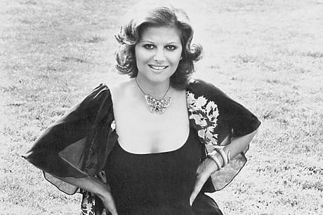 Smiling woman with short hair in a patterned dress seated on grass, wearing a decorative necklace and bracelets.