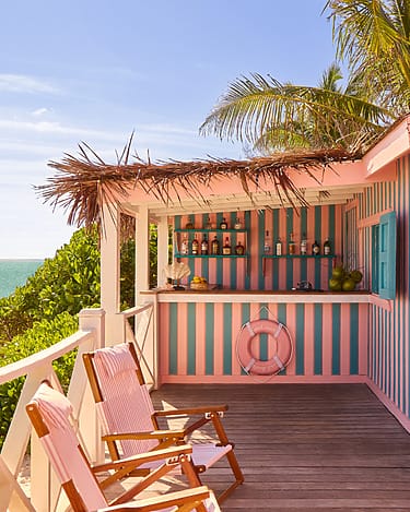 Tropical beach bar with striped pink and teal decor, drinks on counter, two wooden chairs, ocean in background.