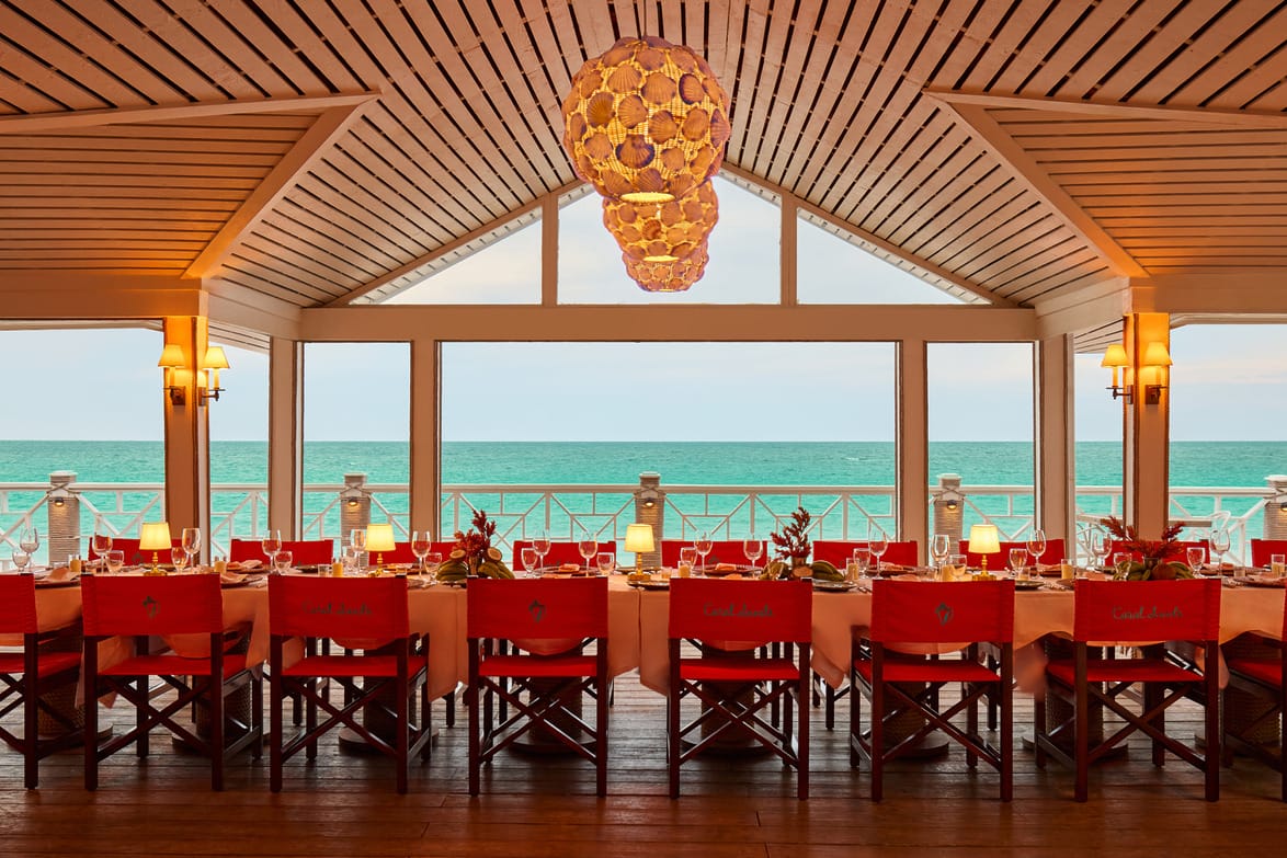 Beachfront restaurant with long dining table set for dinner, overlooking the ocean through large windows under a wooden ceiling.