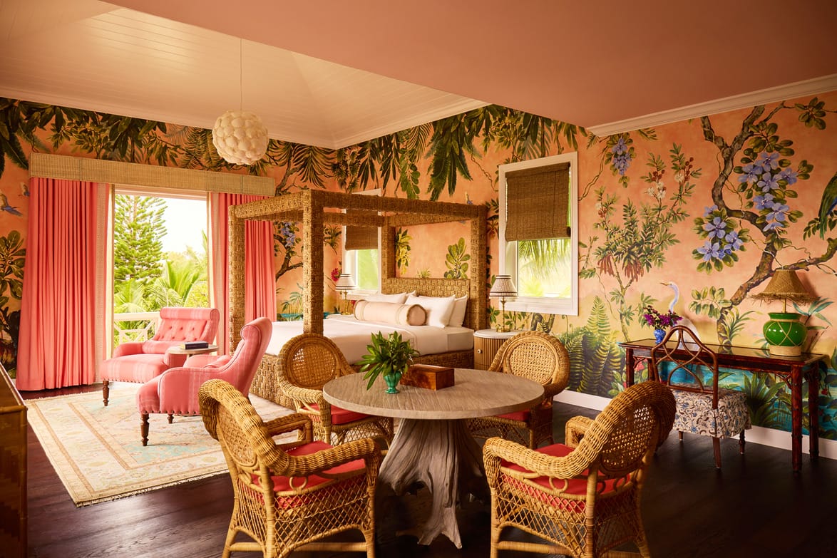 Tropical-themed hotel room with wicker chairs, pink walls, a king-size bed, and lush greenery visible through the window.