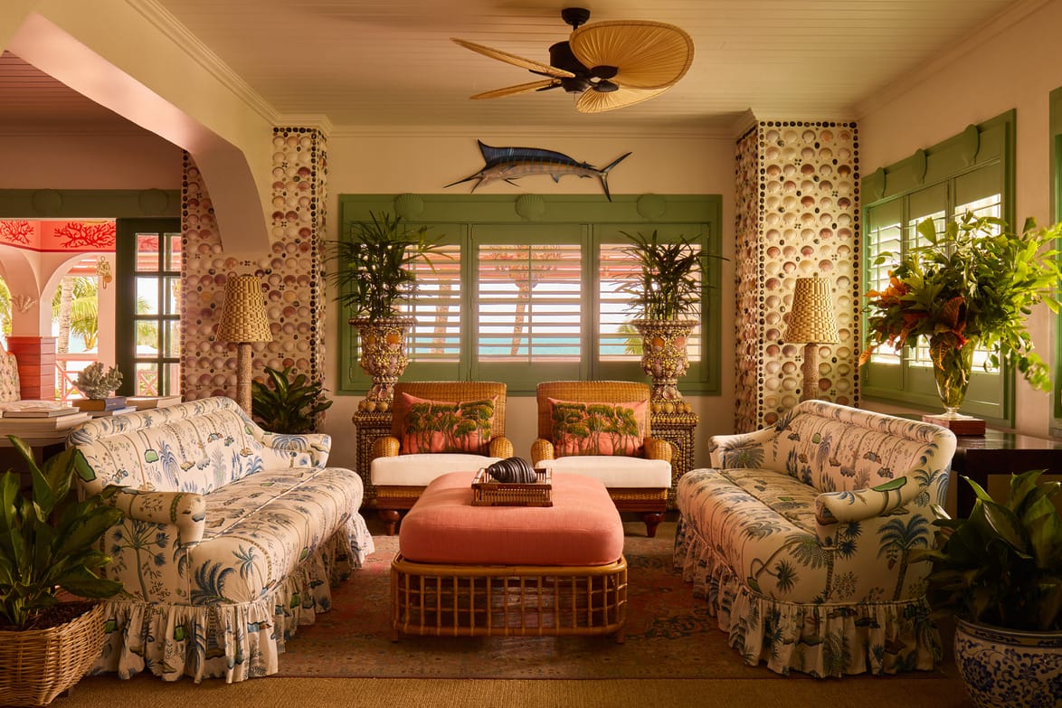 Tropical-themed living room with floral sofas, wicker ottoman, plants, ceiling fan, and a swordfish decoration on the wall.