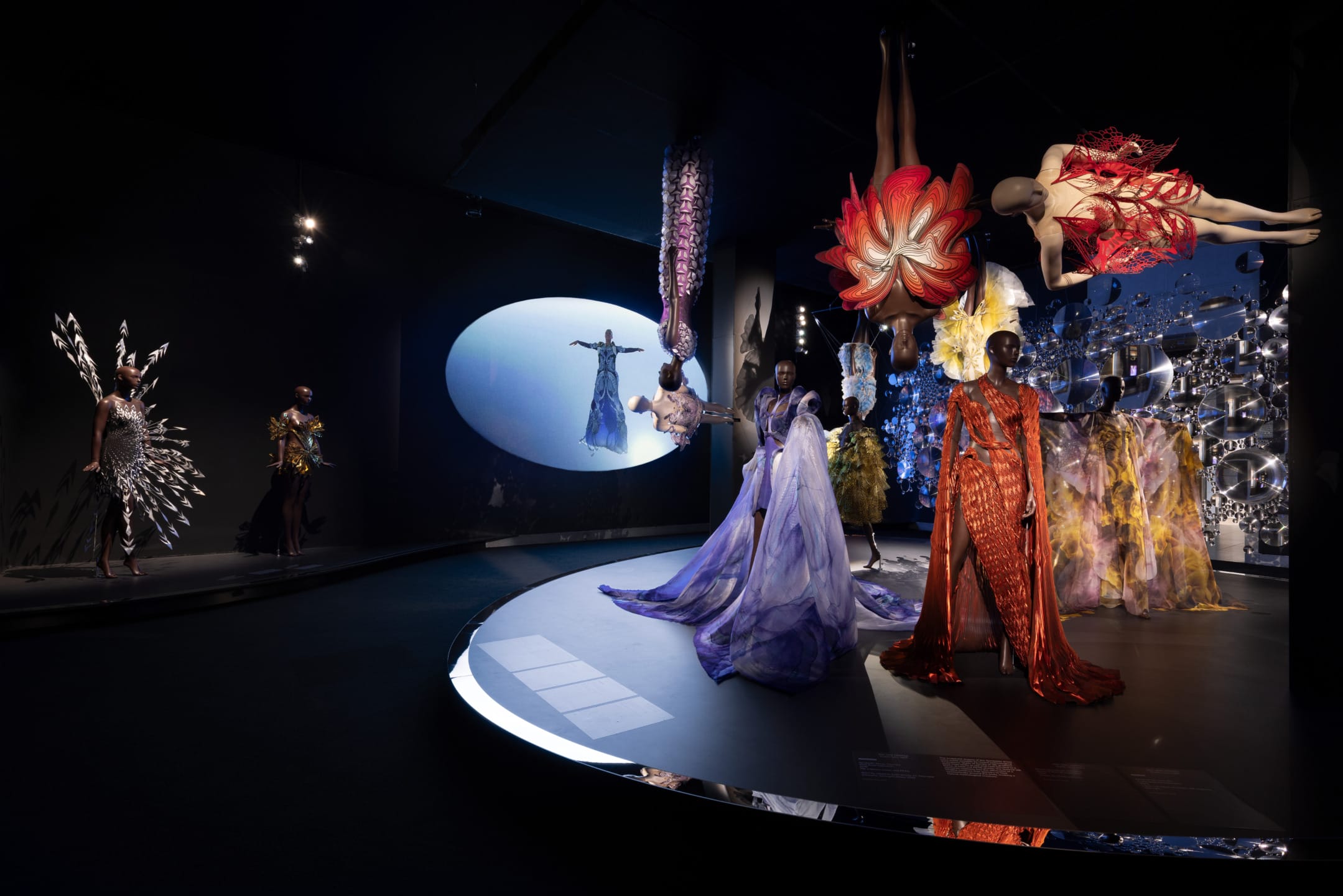 Fashion exhibit with vibrant, avant-garde dresses displayed on mannequins in a dimly lit gallery setting.