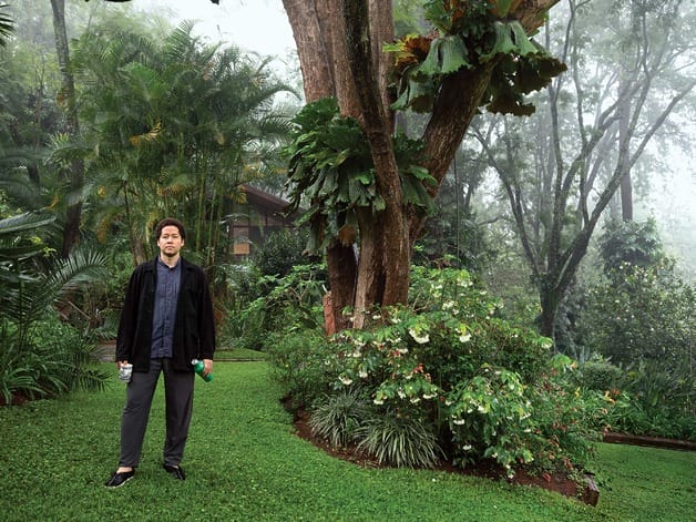 Man in a garden holding a water bottle, surrounded by lush green plants and trees on a misty day.