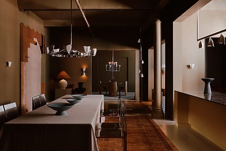 Dimly lit dining room with modern decor, long table, metal chairs, and unique lighting fixtures.