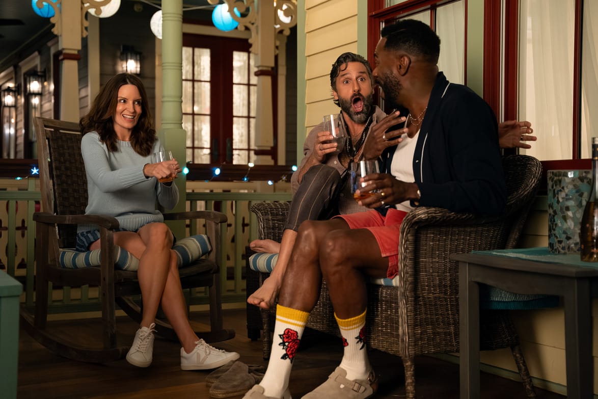 Three people laughing and enjoying drinks on a porch, sitting in wooden chairs, surrounded by warm lighting and decor.
