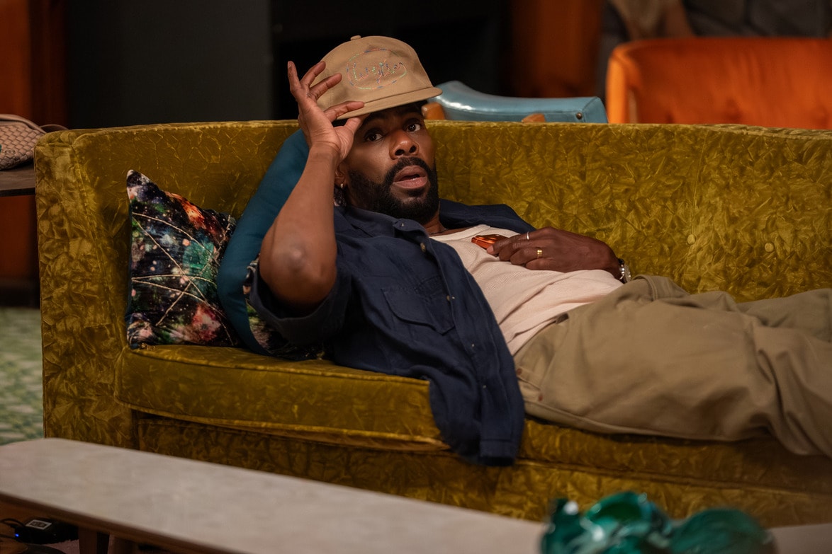Man lying on a green sofa, adjusting his hat while looking intrigued.