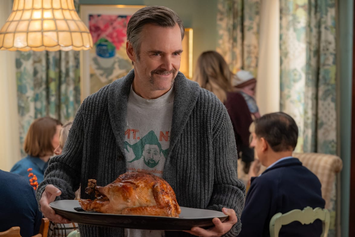 Person holding a roasted turkey on a tray, surrounded by people seated at a dining table in a cozy room.
