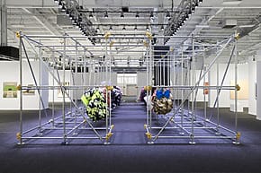 Art installation with geometric metal framework and colorful fabric spheres in a gallery setting with bright overhead lights.