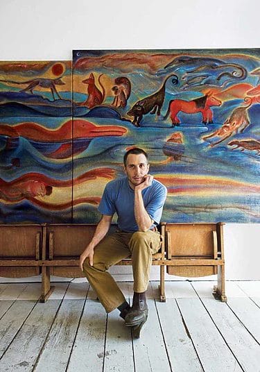 Man sitting on wooden bench in front of colorful animal-themed artwork, white wall and wooden floor in background.