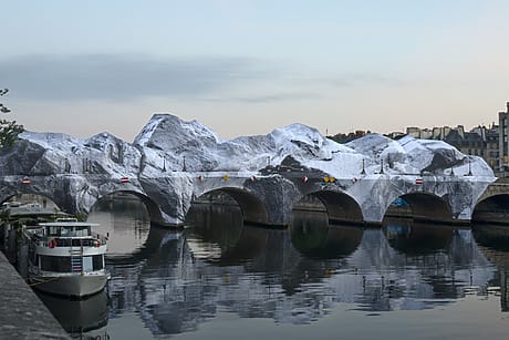 Artistically wrapped bridge resembling snowy mountains, reflected in calm river with parked boat nearby.