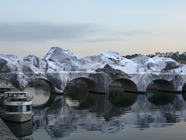 Artistically wrapped bridge resembling snowy mountains, reflected in calm river with parked boat nearby.