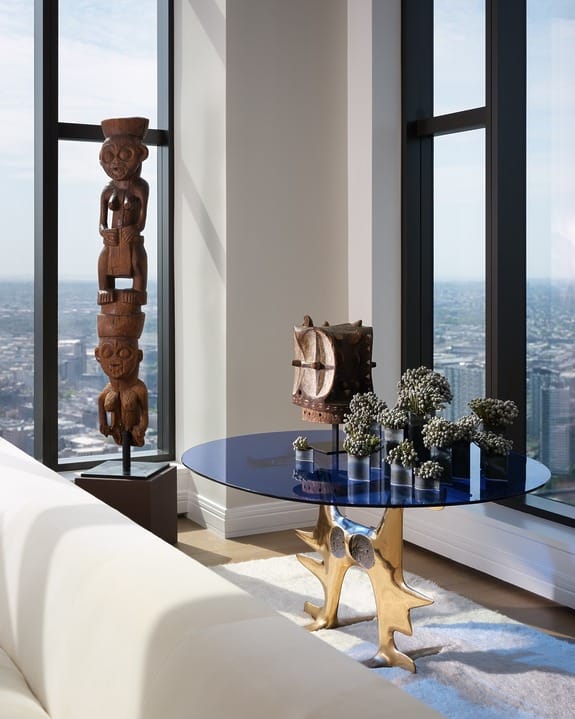 Stylish living room with blue glass table, gold base, tribal art, and city view through large windows.