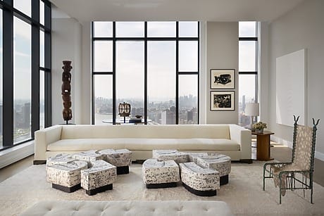 Spacious modern living room with large windows, white sofa, patterned ottomans, and views of the city skyline.