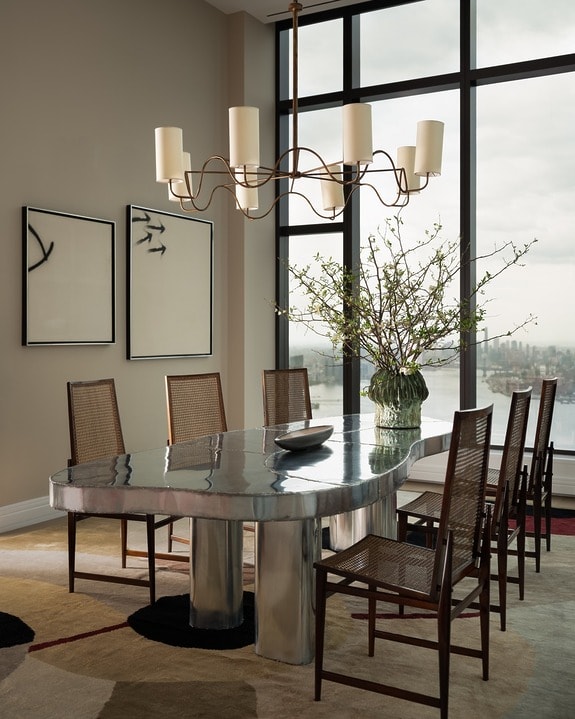 Modern dining room with metallic table, eight chairs, large window view, abstract wall art, and a decorative plant centerpiece.