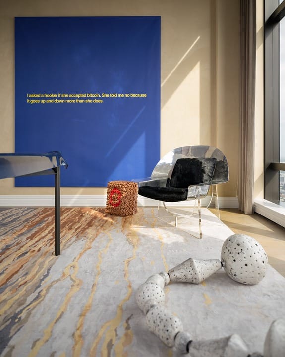 Modern living room with large blue artwork, abstract sculpture, clear chair, woven stool, and floor-to-ceiling windows.