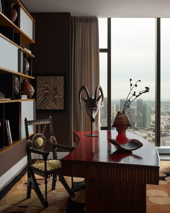 Elegant office with wooden desk, tribal mask decor, and cityscape view from large window. Cozy chair and shelf with books.