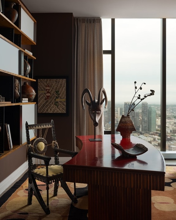 Elegant office with wooden desk, tribal mask decor, and cityscape view from large window. Cozy chair and shelf with books.