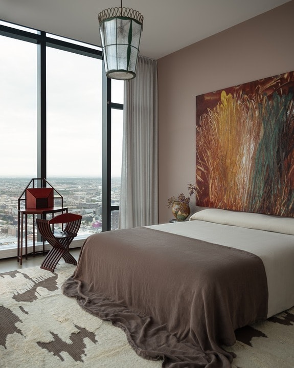 Modern bedroom with large window, abstract painting above bed, and cozy decor, overlooking cityscape.