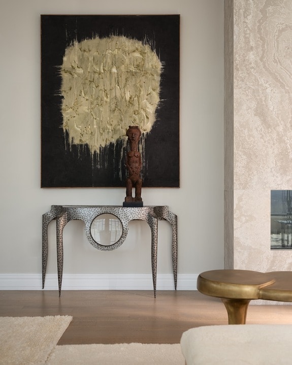 Modern living room with abstract painting, tribal sculpture on a metal console table, neutral tones, and minimalist decor.