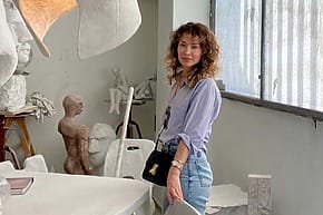 Woman standing in an art studio with sculptures and a sunlit window in the background.