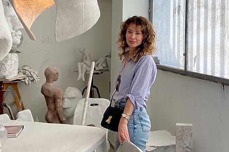 Woman standing in an art studio with sculptures and a sunlit window in the background.