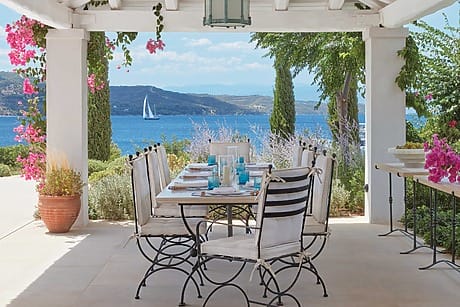Outdoor dining area with sea view, pink flowers, and sailing boat in the distance under a pergola.